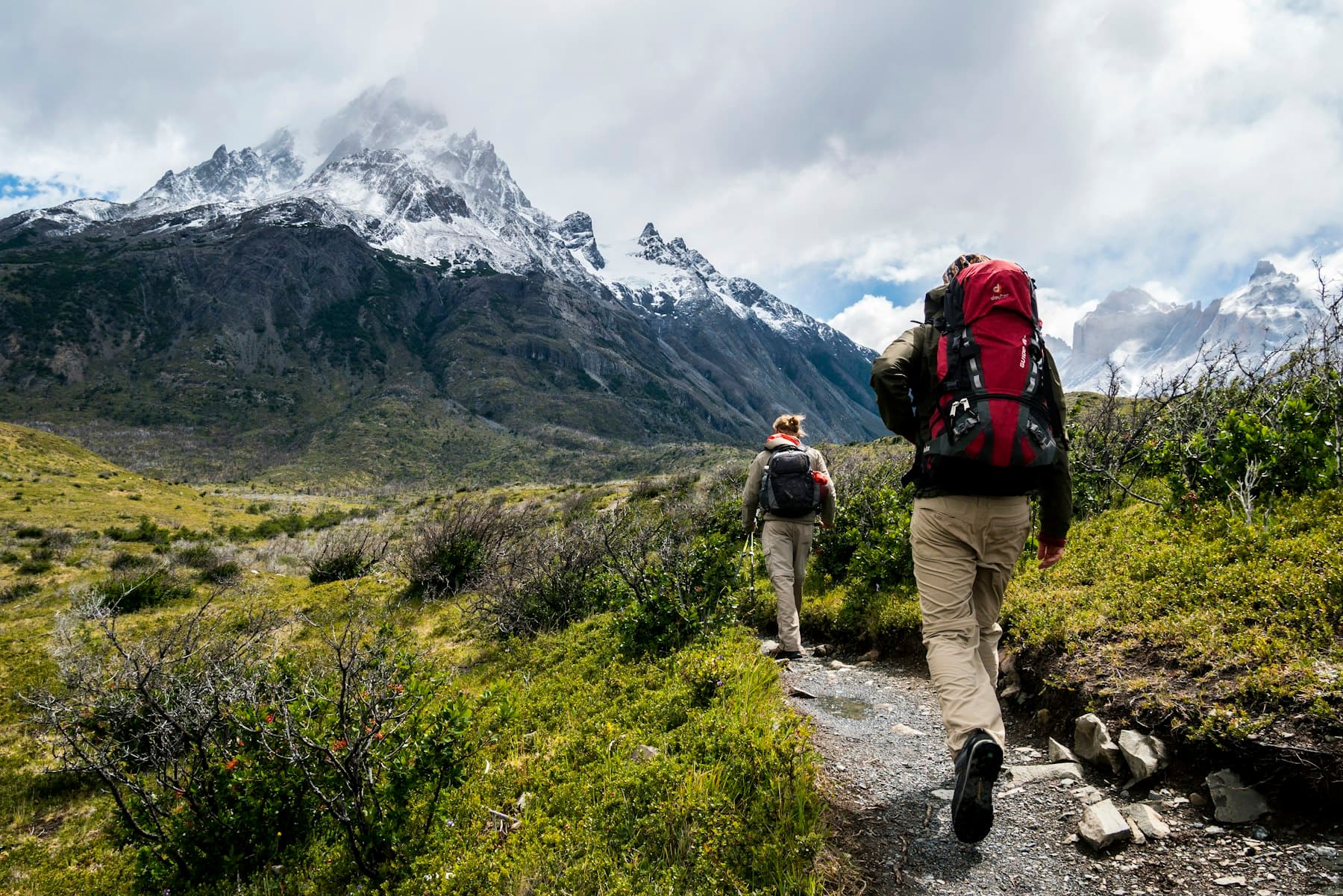 Two walkers on a rocky alpine trail toward a snow-capped peak